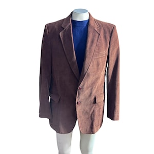 Può includere: Un blazer marrone in velluto a coste con due tasche frontali e due bottoni. Il blazer è esposto su un manichino con un maglione blu a coste sotto. Il blazer ha un classico risvolto.