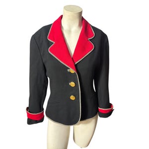 May include: Black blazer with a red collar and cuffs. The blazer has gold buttons and white trim along the edges. The jacket is displayed on a mannequin.