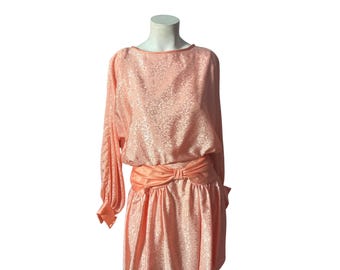 May include: A peach-colored dress with a floral pattern. The dress features long sleeves, a boat neckline, and a cinched waist with a bow detail. The skirt has a gathered design. The dress is displayed on a mannequin.