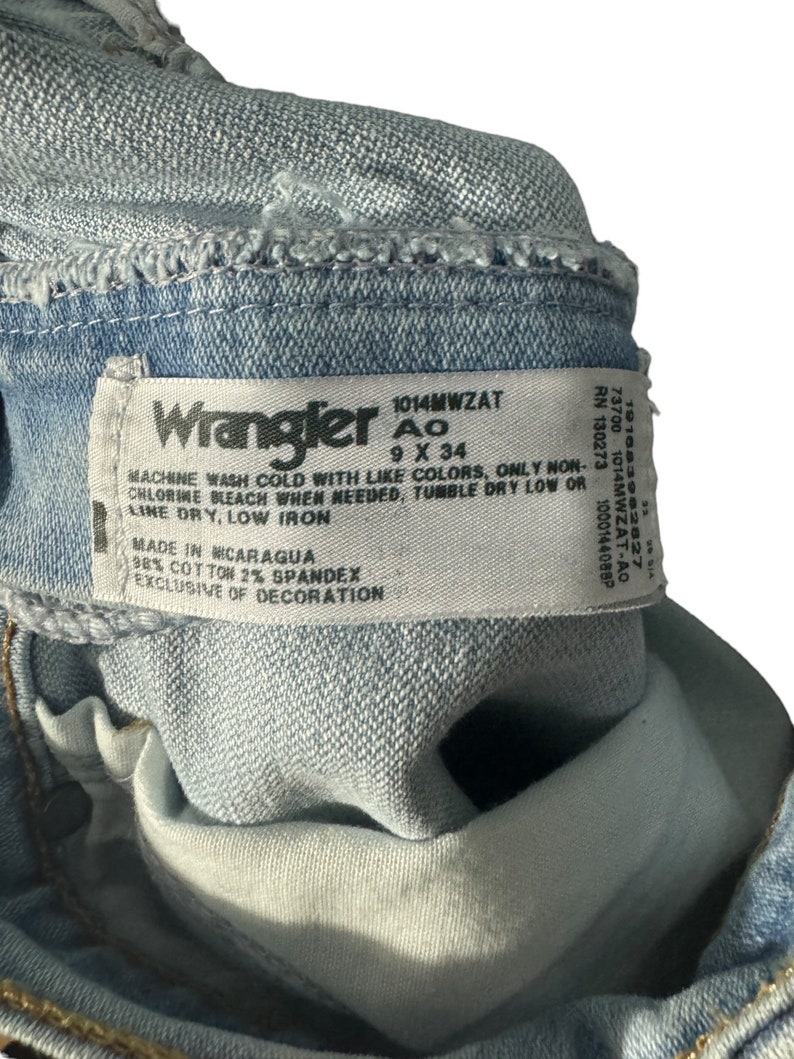 May include: A close-up of a blue denim Wrangler jeans label with the text "1014MWZAT Ao 9 X 34" and care instructions. The label also states "Made in Nicaragua 98% Cotton 2% Spandex Exclusive of Decoration."