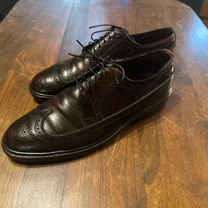 Puede incluir: Un par de zapatos de vestir de cuero negro con diseño de brogue y punta de ala. Los zapatos tienen cierre de cordones y un acabado pulido.