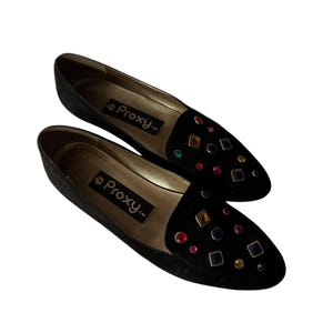 Puede incluir: Par de mocasines de ante negro con adornos de joyas de colores. Los mocasines tienen la palabra "Proxy" impresa en la plantilla.