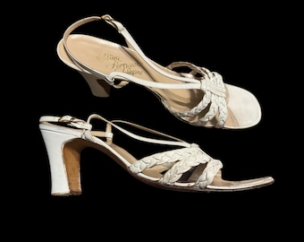 Puede incluir: Dos sandalias de cuero blanco con correas trenzadas y un tacón pequeño. Las sandalias tienen una correa trasera y un cierre de hebilla. El nombre de la marca "Nina Ferragamo" está impreso en la plantilla.