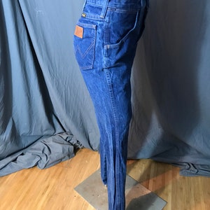 May include: Dark blue denim jeans with a flared leg design. The jeans feature a classic five-pocket style, a brown leather patch on the back, and the Wrangler logo. The jeans are displayed on a mannequin.