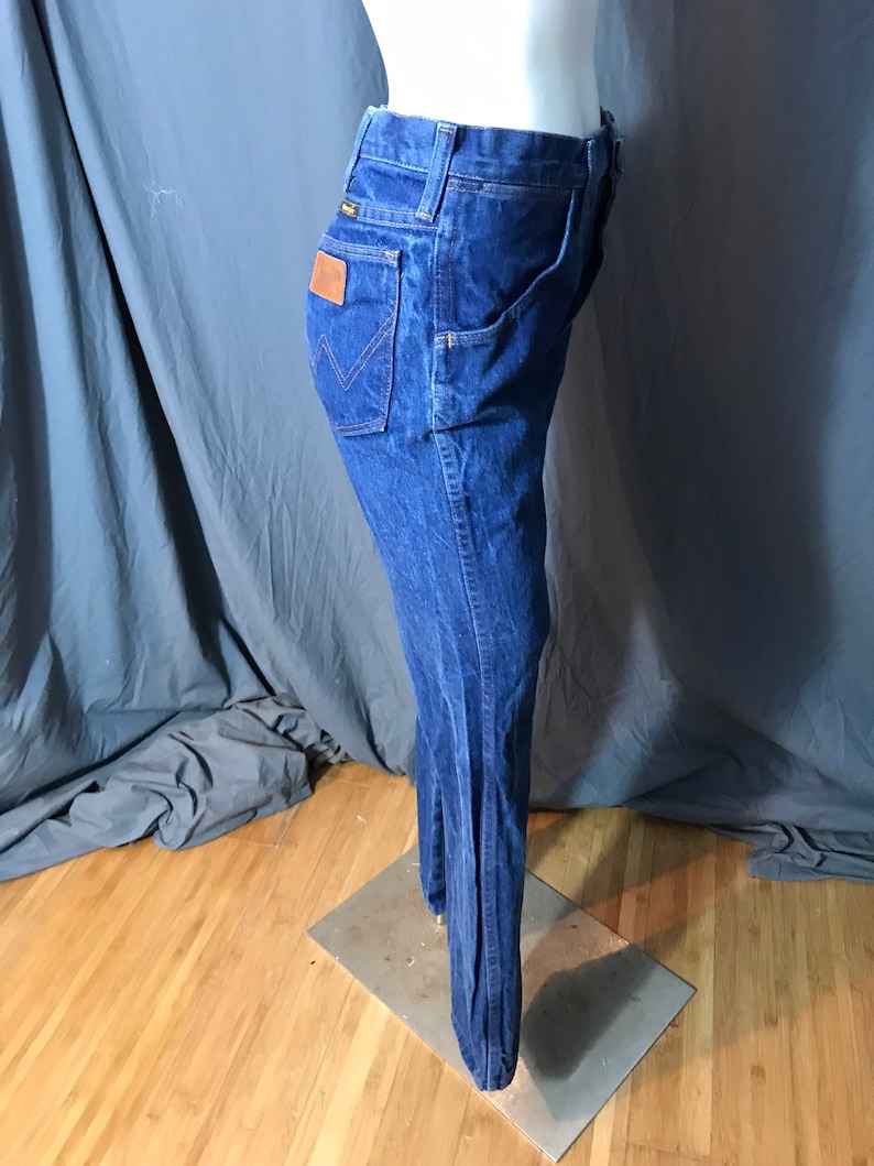 May include: Dark blue denim jeans with a flared leg design. The jeans feature a classic five-pocket style, a brown leather patch on the back, and the Wrangler logo. The jeans are displayed on a mannequin.