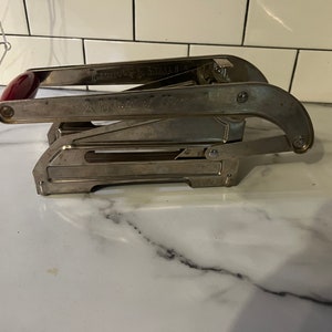May include: A vintage, silver-toned can opener with a red handle. The can opener has the text "A. West & Co." and "Improved Can Opener" etched on the metal. The can opener is on a white marble surface.