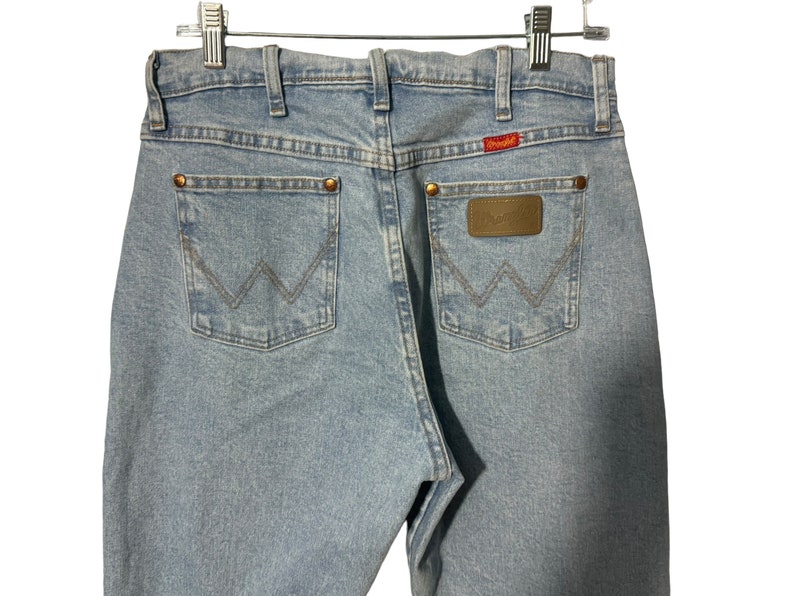 May include: Light blue denim jeans with a brown leather patch on the back pocket that says "Wrangler".