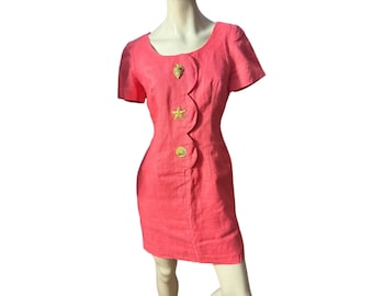 Könnte beinhalten: Ein korallenfarbenes Kleid mit rundem Ausschnitt und kurzen Ärmeln. Das Kleid hat ein dekoratives, wellenförmiges Design auf der Vorderseite, verziert mit goldfarbenen Muschel-, Seestern- und Muschel-Verzierungen. Das Kleid wird auf einer Schaufensterpuppe präsentiert.