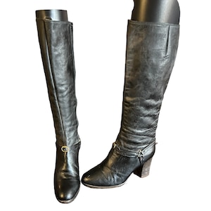 Peut inclure: Bottines noires en cuir montant au genou avec une boucle dorée et un talon bloc.