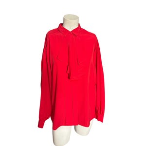 May include: A vibrant red blouse with long sleeves and a collared neckline. The blouse features a unique bow detail at the neck and is displayed on a mannequin. The fabric appears to be smooth and flowing.