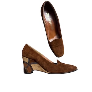 May include: Two brown suede wedge heels. One shoe is positioned above the other. The wedge heel features a geometric design in shades of brown and tan. The shoes have a pointed toe and a low-cut vamp.