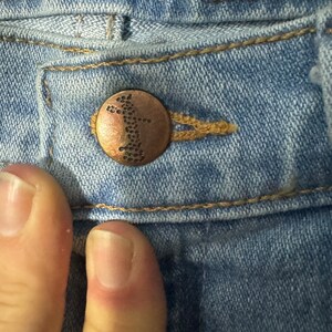 May include: Close-up of a light blue denim jean with a copper-coloured button. The button has a small, intricate design.