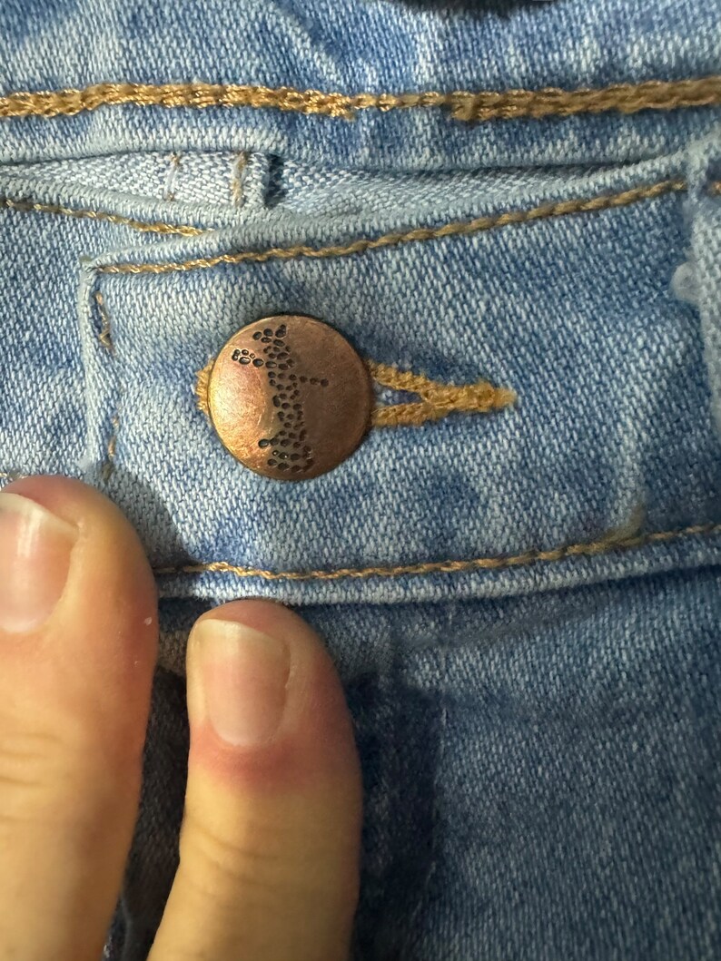 May include: Close-up of a light blue denim jean with a copper-coloured button. The button has a small, intricate design.