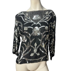 Könnte beinhalten: Ein schwarzes Paillettenoberteil mit silbernem Blumendesign. Das Oberteil hat einen Boot Ausschnitt und Dreiviertelärmel.