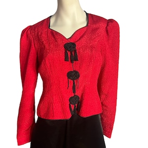 Może przedstawiać: Czerwona, fakturowana, krótka kurtka z czarnymi guzikami z frędzlami. Kurtka ma lekko bufiasty rękaw i dopasowany pas.