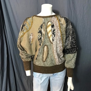 Può includere: Un maglione marrone, beige e grigio con un motivo geometrico. Il maglione ha un collo rotondo e maniche lunghe.