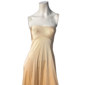 May include: A strapless, floor-length, champagne-colored slip dress. The dress is displayed on a mannequin. The fabric appears to be a soft, silky material. The bodice has a subtle V-neck design, and the skirt flows smoothly.