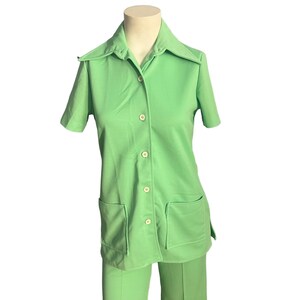 May include: A light green two-piece outfit, consisting of a short-sleeved button-down shirt with two front pockets and matching pants. The outfit is displayed on a mannequin. The shirt has a wide collar and is buttoned up with four buttons.