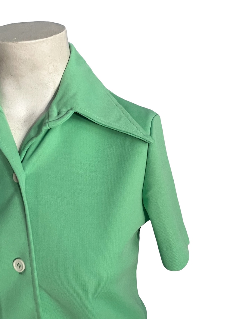 May include: A light green short-sleeved shirt with a pointed collar and two white buttons. The shirt is made of a smooth fabric and has a classic design. The shirt is displayed on a mannequin.