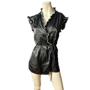 Op de afbeelding: Zwarte satijnen wikkel top met witte bies en bijpassende strikriem. De top heeft gefranjerde kapmouwen en een gefranjerde kraag. Het kledingstuk wordt op een mannequin gepresenteerd.