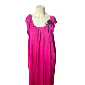 May include: A bright pink, floor-length nightgown with short sleeves and a scoop neckline. A black bow is attached to the left shoulder. The gown is displayed on a mannequin.