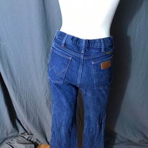 May include: Dark blue denim jeans with a classic fit. The jeans feature a brown leather patch on the back pocket and the Wrangler logo. The jeans are displayed on a mannequin against a gray backdrop.