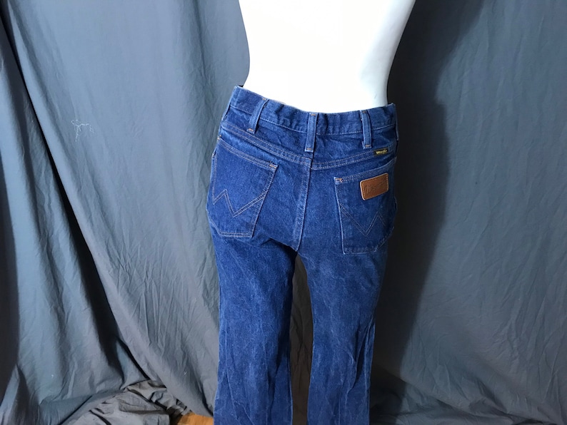 May include: Dark blue denim jeans with a classic fit. The jeans feature a brown leather patch on the back pocket and the Wrangler logo. The jeans are displayed on a mannequin against a gray backdrop.