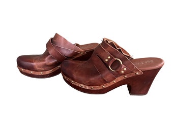 May include: Brown leather clogs with a buckle and studded platform. The clogs have a wooden heel and are made by the brand "Kalin"