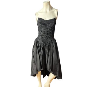 May include: A black strapless dress with a ruched bodice and a high-low skirt. The dress is displayed on a mannequin. The fabric has a slight sheen, suggesting a satin or similar material. The dress is a formal or semi-formal style.