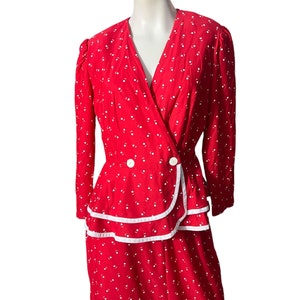 Puede incluir: Un vestido rojo con lunares blancos y un ribete blanco alrededor del escote y el dobladillo. El vestido tiene un corpiño estilo envolvente y una falda con un volante de capas.