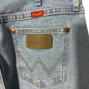 May include: Light blue denim jeans with a brown leather Wrangler patch on the back pocket. The patch features the Wrangler logo in white lettering.