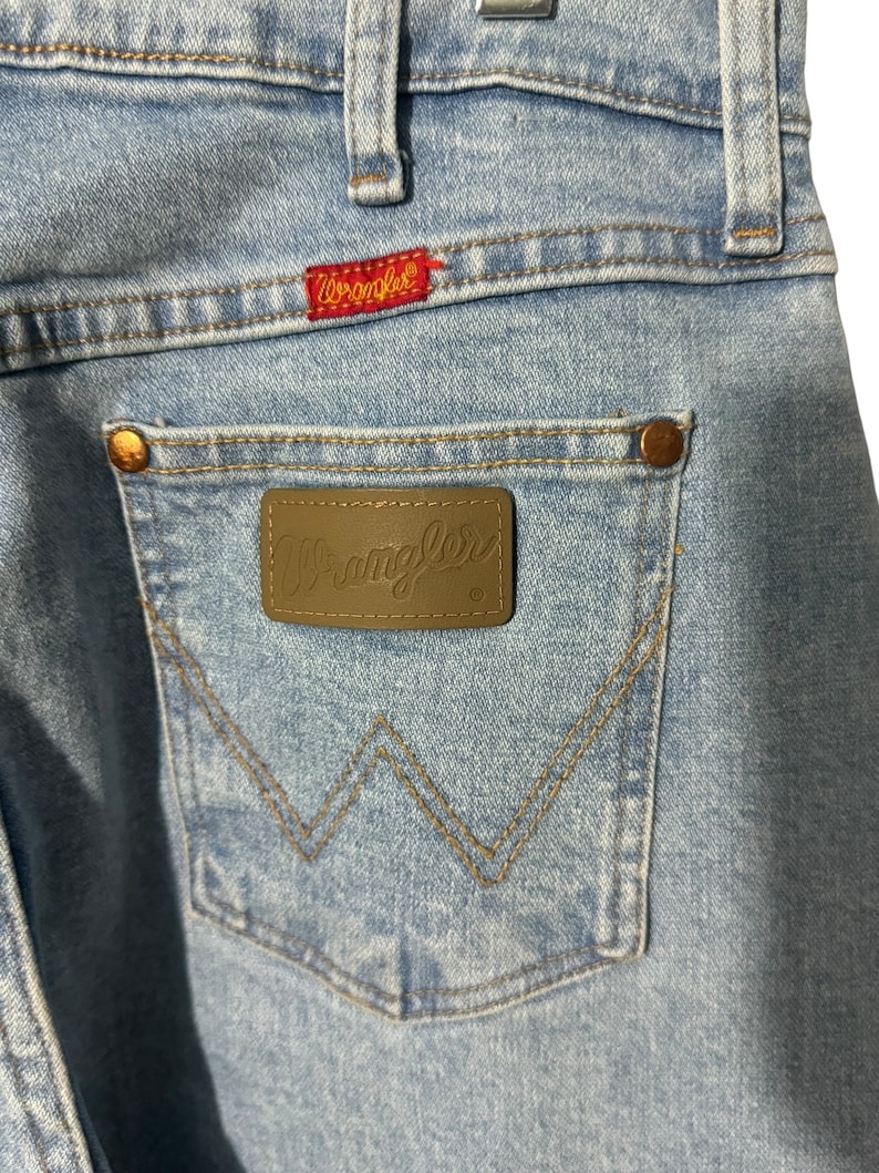 May include: Light blue denim jeans with a brown leather Wrangler patch on the back pocket. The patch features the Wrangler logo in white lettering.