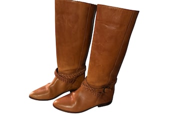 Peut inclure: Une paire de bottes en cuir marron montant au genou avec un détail de sangle tressée.