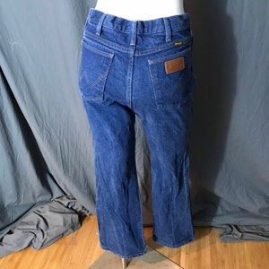 May include: Blue denim Wrangler jeans with a classic five-pocket design. The jeans feature a brown leather patch with the Wrangler logo on the back pocket. The jeans are displayed on a mannequin.