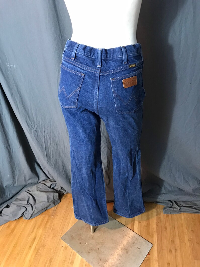 May include: Blue denim Wrangler jeans with a classic five-pocket design. The jeans feature a brown leather patch with the Wrangler logo on the back pocket. The jeans are displayed on a mannequin.