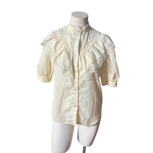 May include: Cream-colored blouse with a high collar and short sleeves. The blouse features a button-down front and decorative lace detailing on the collar and shoulders. The fabric appears lightweight and slightly sheer.