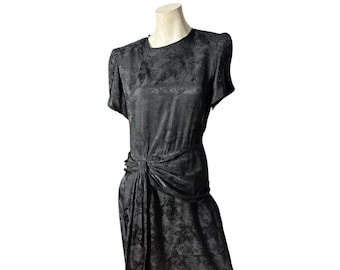 May include: A black vintage dress with a floral pattern, displayed on a mannequin. The dress features short sleeves, a gathered waist, and a draped detail on the skirt. The fabric has a subtle sheen.
