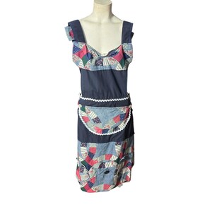 Op de afbeelding: Een vintage patchwork schortjurk met een marineblauwe lijfje en bandjes. De jurk heeft een top in bib-stijl met een patchwork ontwerp van rode, blauwe en groene stoffen. De schort heeft een witte geschulpte rand en een patchwork zak.