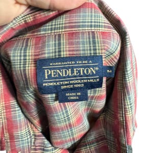 Op de afbeelding: Een rood, grijs en beige geruit overhemd met een Pendleton-label. Het label luidt "Warranted to be a Pendleton Woolen Mills Since 1863 Made in China". Het overhemd is maat M.