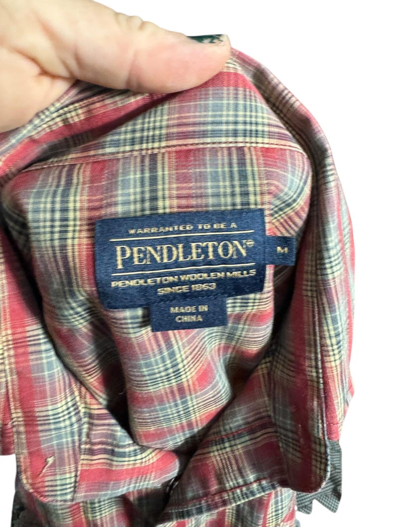 Op de afbeelding: Een rood, grijs en beige geruit overhemd met een Pendleton-label. Het label luidt "Warranted to be a Pendleton Woolen Mills Since 1863 Made in China". Het overhemd is maat M.