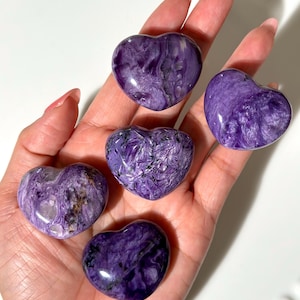 Puede incluir: Se muestran cinco piedras pulidas en forma de corazón en tonos morados y blancos. Las piedras tienen un aspecto veteado y se sostienen en una mano abierta. El fondo es una superficie blanca brillante.