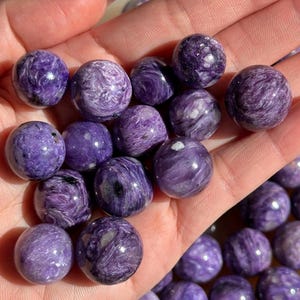 May include: A collection of polished, spherical gemstones in varying shades of purple, with swirling patterns and a glossy finish. The stones are held in a hand, showcasing their smooth texture and natural beauty. These stones are approximately 2.5 cm in diameter.