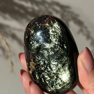 May include: A polished, oval gemstone with a dark green and gold iridescent sheen. The stone has a smooth, rounded form and reflects light, showcasing a swirling pattern. The gemstone is held in a hand, with a neutral background.
