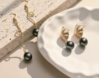 Könnte beinhalten: Goldfarbene Ohrringe mit dunkelgrauen Perltropfen. Ein Paar hat ein langes, geschwungenes Design, das andere hat einen muschelartigen Aufsatz. Die Ohrringe werden auf einem weißen, wellenförmigen Teller und einer strukturierten Oberfläche präsentiert.