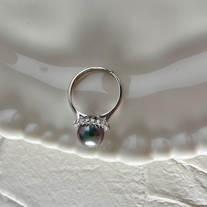 K&ouml;nnte beinhalten: Ein silberner Ring mit einer irisierenden Perle. Die Perle hat eine dunkle, mehrfarbige Oberfl&auml;che. Die Fassung des Rings ist mit kleinen, klaren Edelsteinen verziert. Der Ring wird auf einem wei&szlig;en, wellenf&ouml;rmigen Teller pr&auml;sentiert.