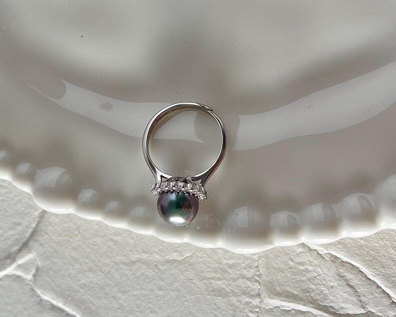 K&ouml;nnte beinhalten: Ein silberner Ring mit einer irisierenden Perle. Die Perle hat eine dunkle, mehrfarbige Oberfl&auml;che. Die Fassung des Rings ist mit kleinen, klaren Edelsteinen verziert. Der Ring wird auf einem wei&szlig;en, wellenf&ouml;rmigen Teller pr&auml;sentiert.