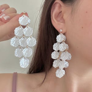 May include: White, layered chandelier earrings with a pearlescent finish. Each earring features multiple tiers of petal-shaped elements, connected by delicate gold-colored links. A small, clear gem adorns the top of each earring.