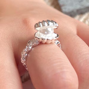 May include: A silver-colored ring featuring a seashell design with a pearl. The ring's band has a textured, ornate pattern. The seashell opens to reveal a white pearl, creating a unique and elegant piece of jewelry.