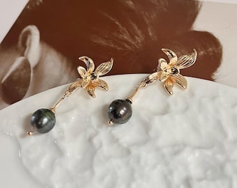 Könnte beinhalten: Goldfarbene Blumenohrringe mit einer hängenden dunkelgrauen Perle. Die Ohrringe werden auf einer weißen, strukturierten Oberfläche präsentiert, mit einer teilweise sichtbaren Zeitschrift im Hintergrund. Der Titel der Zeitschrift lautet "the gentlewor".