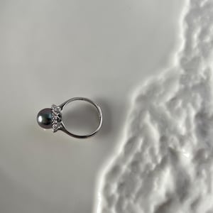 K&ouml;nnte beinhalten: Ein silberner Ring mit einem dunkelgrauen Perlmutt-Mittelstein und einem Kranz aus kleinen, klaren Edelsteinen. Der Ring liegt auf einer wei&szlig;en, strukturierten Oberfl&auml;che, die einen Kontrast erzeugt. Schmuck.